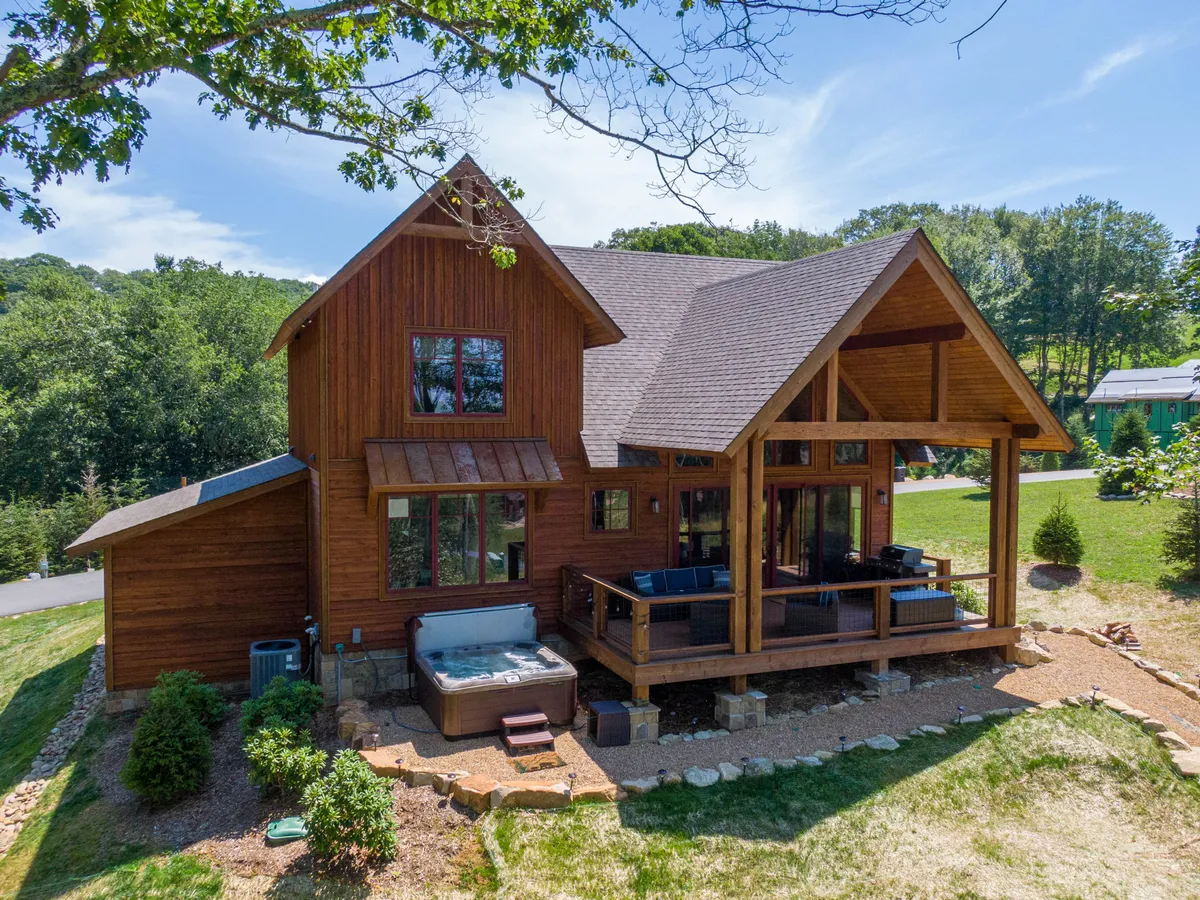 A welcoming wooden house with a spacious deck outfitted with seating and a hot tub. The lush greenery surrounding the deck offers privacy, while the hot tub commands a panoramic view of the mountains. The scene radiates relaxation, inviting guests to unwind outdoors day or night.