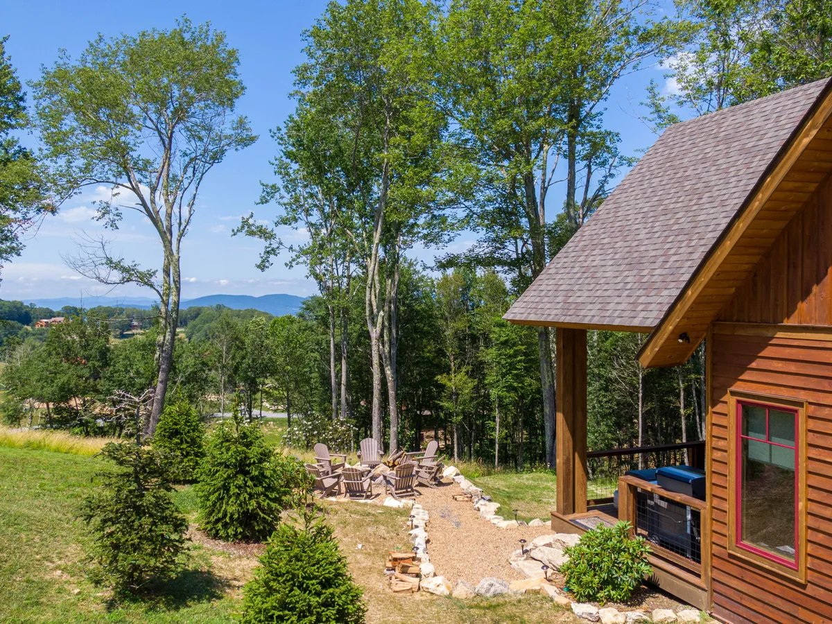 A tranquil exterior view of a wooden cabin nestled among trees, with a seating area, pathway, and a scenic mountain backdrop. The space invites relaxation and outdoor gatherings, ideal for enjoying the natural setting. The image also suggests opportunities for sunrise watching, stargazing, and exploring the immediate environment.