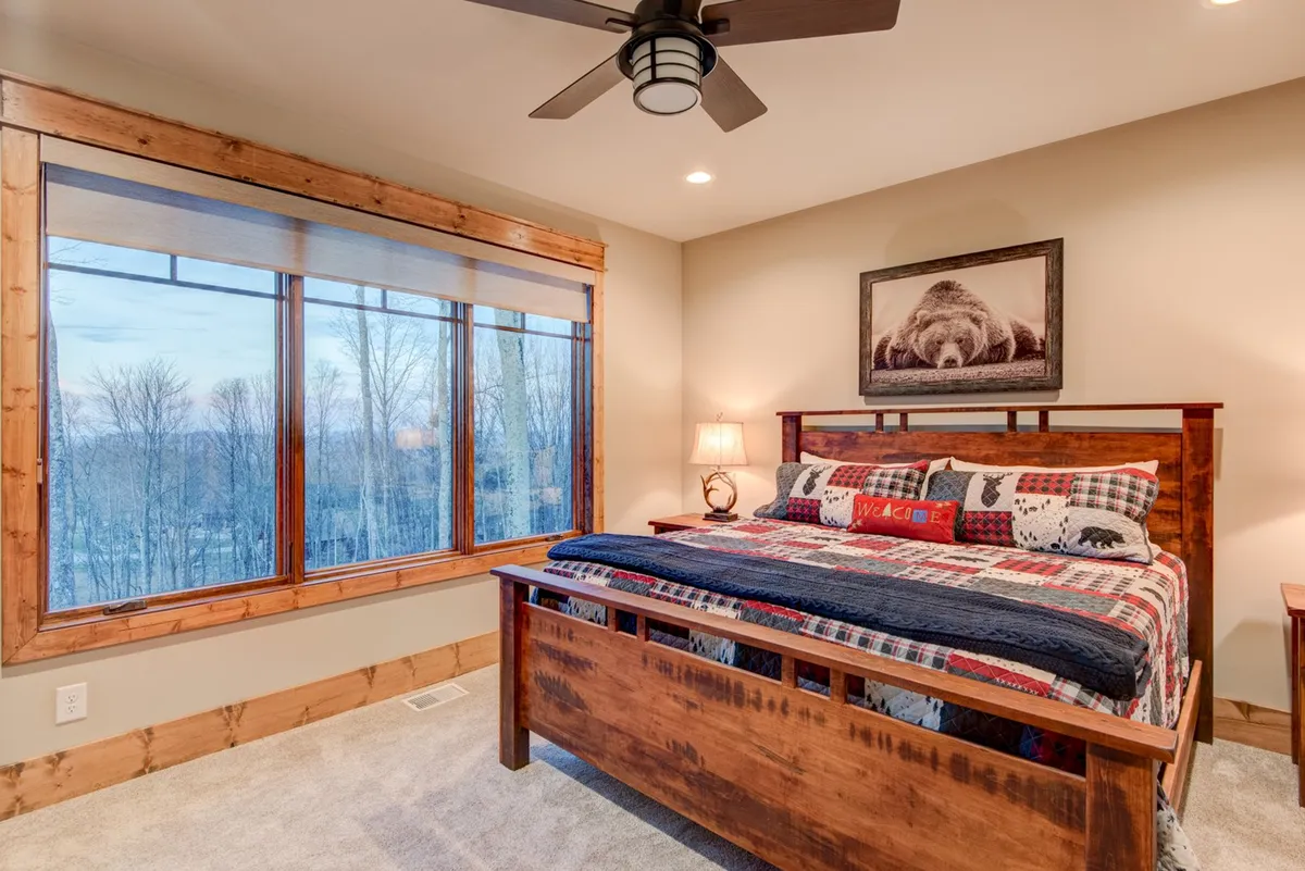 This image captures a bedroom featuring a large window with a beautiful view, a wooden bed frame, colorful bedding, and bear-themed artwork. The inviting, warm atmosphere suggests comfort and relaxation, ideal for restful nights and peaceful mornings.
