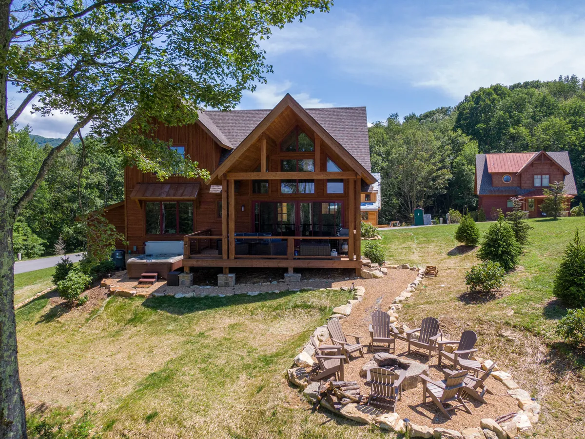 The image shows a charming wooden deck and a cozy fire pit area surrounded by tall trees, with sweeping mountain views in the background. The outdoor space invites guests to relax, dine al fresco, and gather for stories under the stars, all set against the backdrop of nature's tranquility.