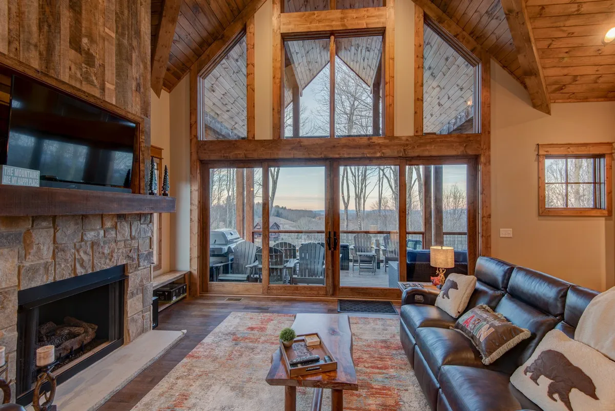 A cozy living room featuring a large leather sofa, a decorative rug, and a stone fireplace, all set against a backdrop of expansive windows with scenic mountain views. The arrangement is ideal for relaxing, watching TV, or gathering with family and friends.
