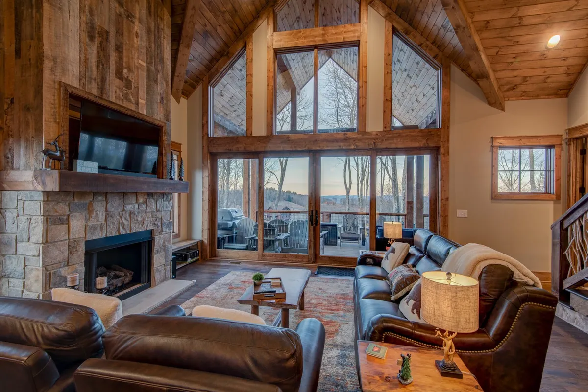 A welcoming living area with large windows overlooking a scenic mountain backdrop, a rustic stone fireplace, comfortable seating, and exposed wooden beams. The space is designed for relaxing, entertaining, or enjoying cozy evenings.