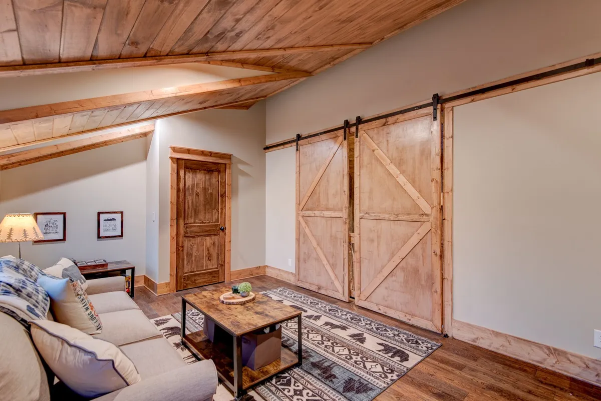 This inviting loft features a comfortable sofa, coffee table set for gathering, decorative rug, table lamp, and striking barn doors. The layout emphasizes the room's flexibility, with ample space for board games or relaxing family time. The barn doors reinforce the idea of privacy when needed.