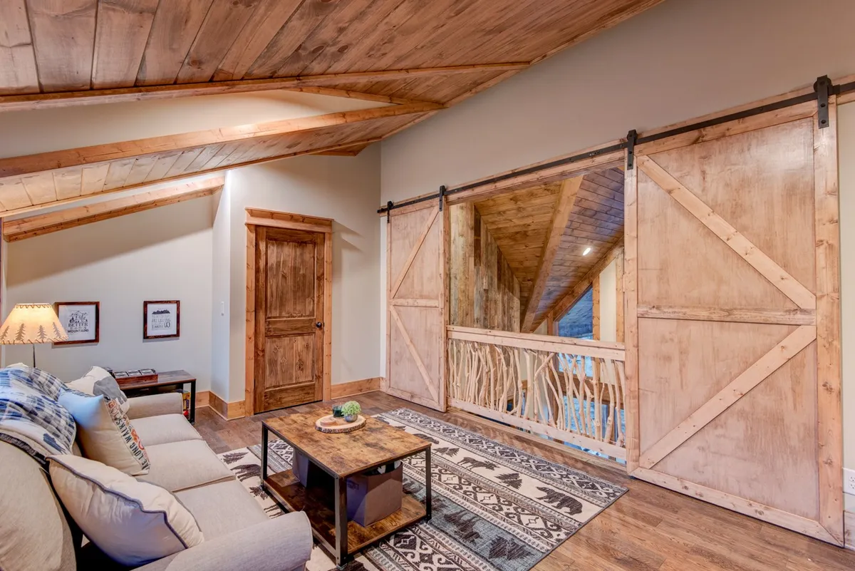 The image features a cozy loft-style interior with a sofa, coffee table, sliding barn doors, and a warm area rug. The space is brightened by natural light and has a welcoming, multi-purpose feel ideal for relaxation, play, or socializing. The sliding barn doors provide a sense of privacy and separation from the rest of the home.