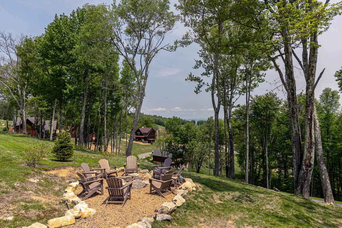 A tranquil fire pit area ringed by Adirondack chairs, set amid tall trees and with distant cabins visible. The ambiance is relaxed, perfect for stargazing or gathering with friends and family under a sunset sky.